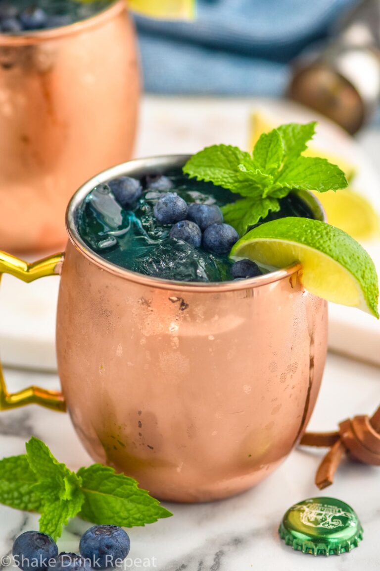 two copper mugs of blue mules with ice, blueberries, lime wedge, and mint leaves