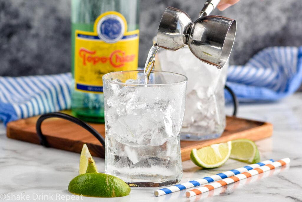 jigger of tequila pouring into a glass of ice to make a Ranch Water cocktail surrounded by fresh lime slices, straws, and Topo Chico mineral water