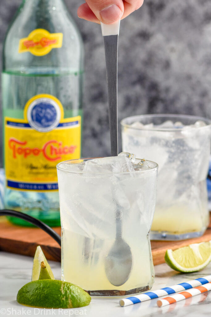 man stirring glass of Ranch Water recipe with ice surrounded by fresh lime wedges and bottle of Topo Chico