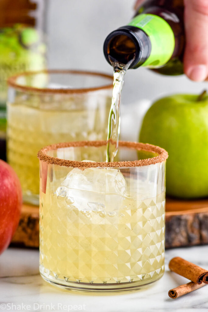 man's hand pouring bottle of hard cider into a glass of apple cider margarita with ice and cinnamon sugar rim. Glass of apple cider margaritas, apples, cinnamon sticks, and bottle of angry orchard hard cider sit in background