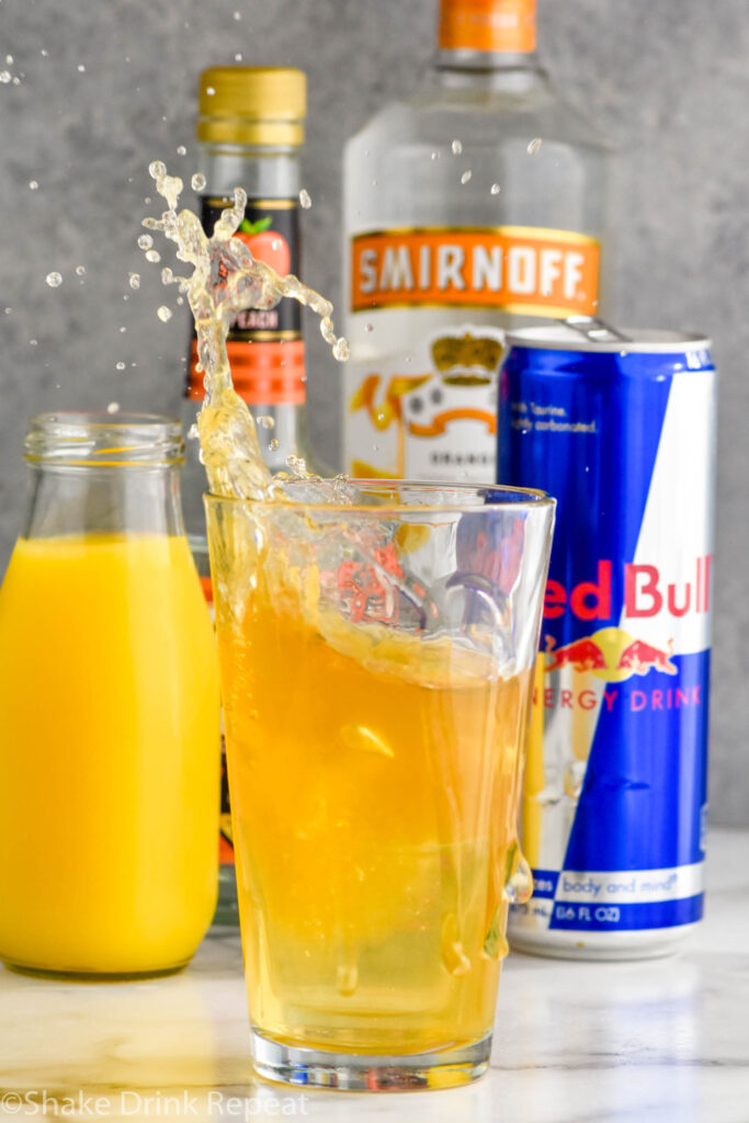 Side view of shot glass being dropped into pint glass of ingredients for cactus cooler shot recipe and making a splash. Bottle of orange juice, peach schnapps, mandarin vodka, and a can of red bull behind glass.