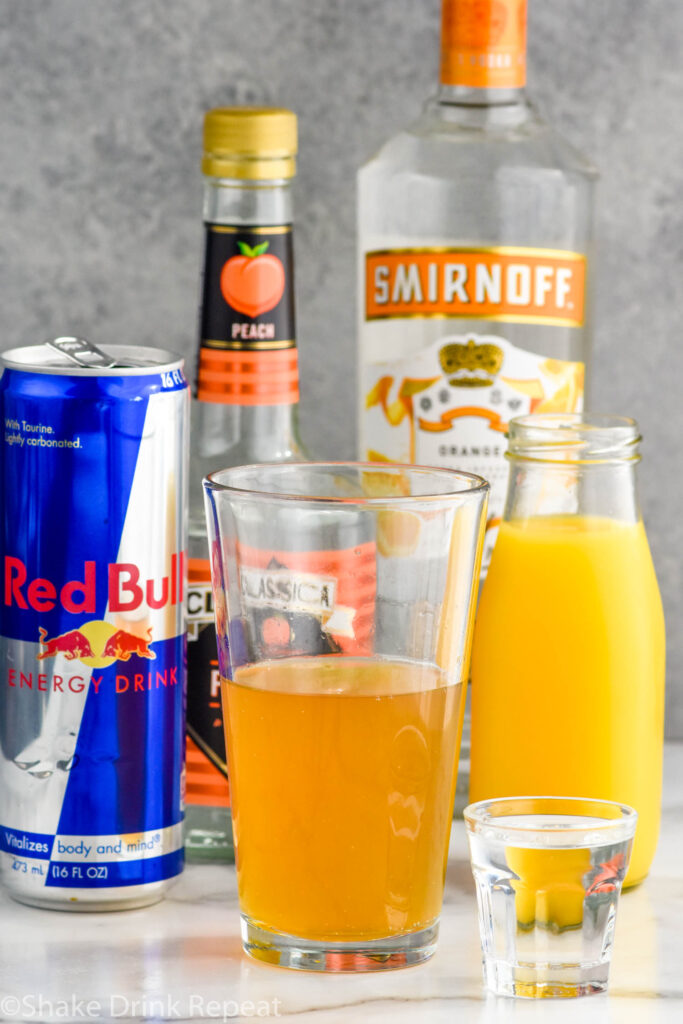 Side view of a pint glass and a shot glass of ingredients for cactus cooler shot recipe. Bottle of orange juice, peach schnapps, mandarin vodka, and a can of red bull.