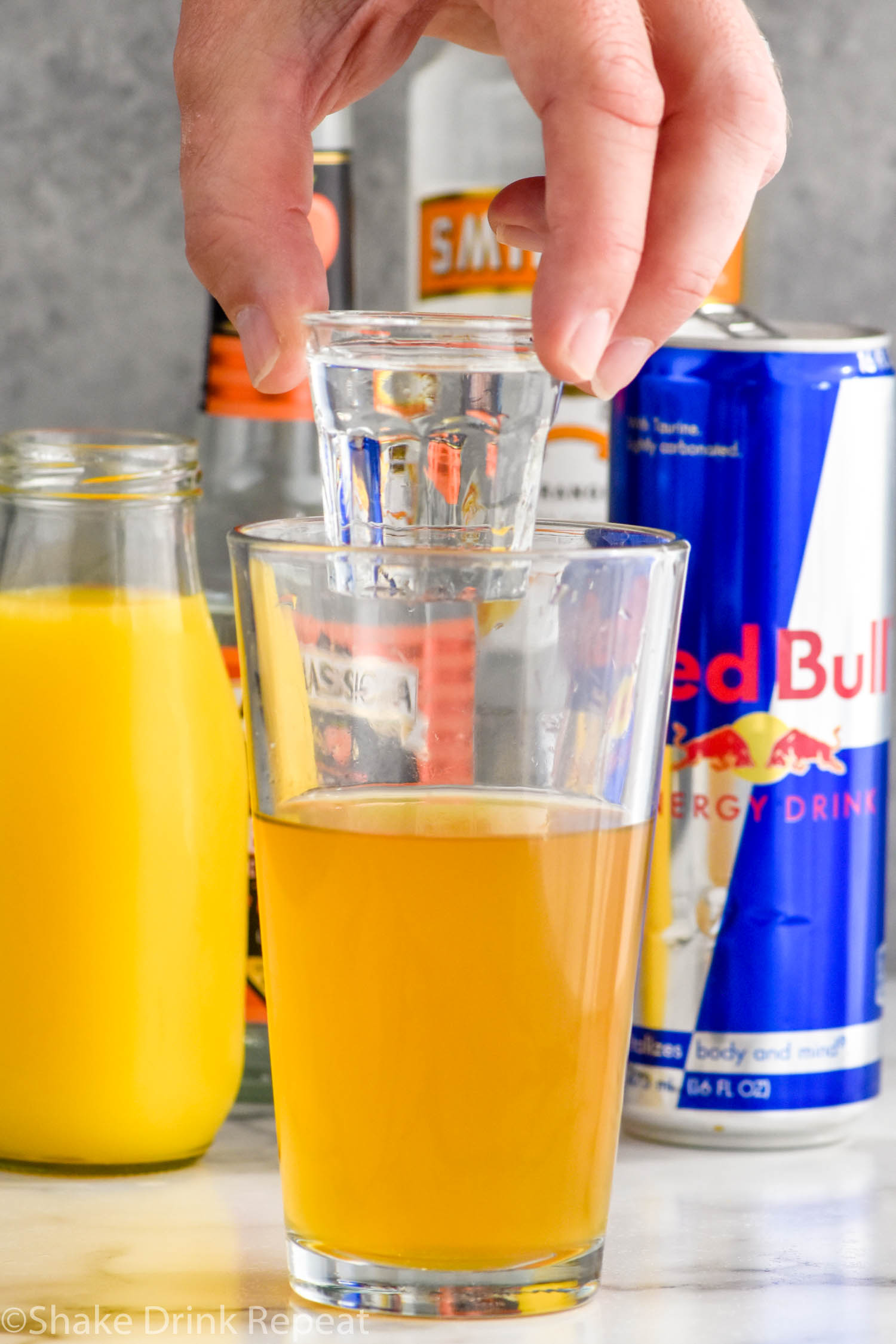Side view of person's hand holding a shot of liqueur over a pint glass of ingredients for Cactus Cooler Shot recipe. Bottle of orange juice, peach schnapps, mandarin vodka, and a can of red bull behind glass.