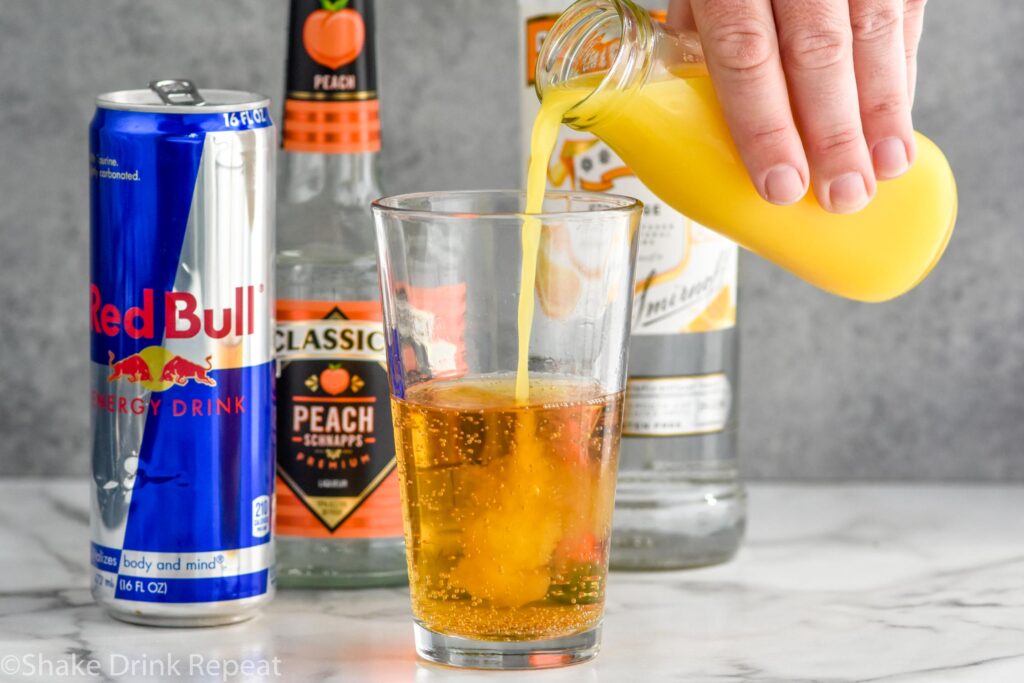 Side view of person's hand pouring orange juice into a pint glass of Red Bull for Cactus Cooler Shot recipe. Behind glass is can of Red Bull, bottle of peach schnapps, and bottle of mandarin vodka.