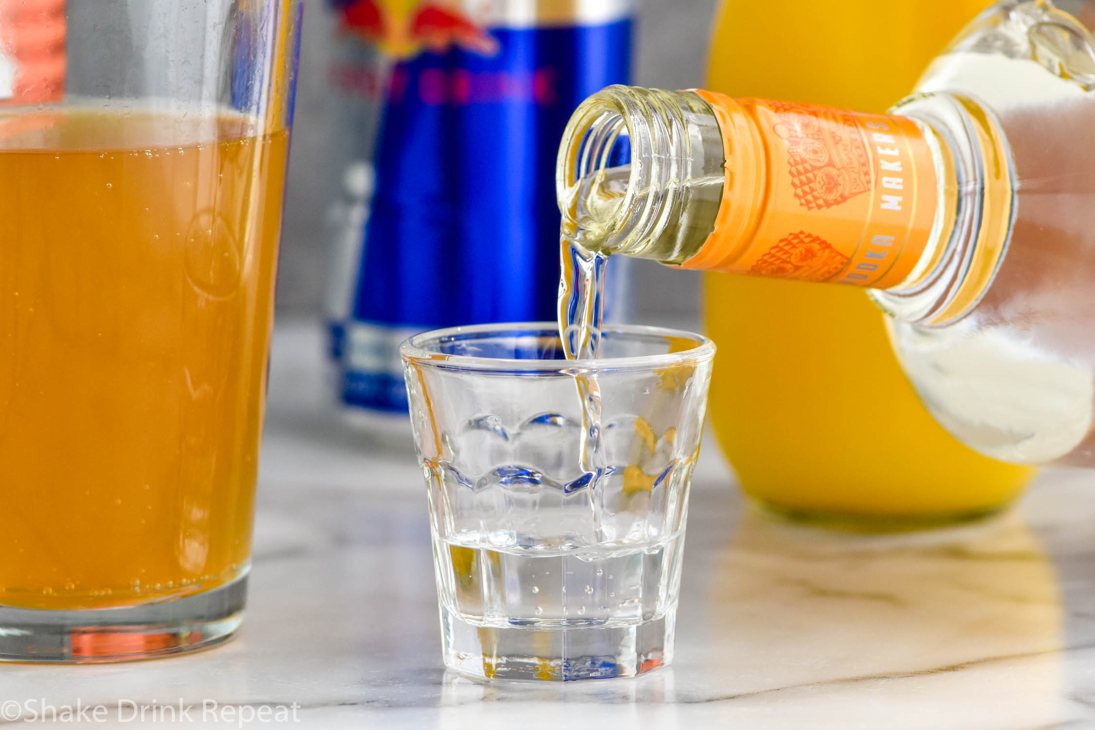 Side view of bottle of mandarin vodka being poured into a shot glass for Cactus Cooler Shot recipe. Pint glass of Red Bull, Red Bull can, and bottle of orange juice in the background.
