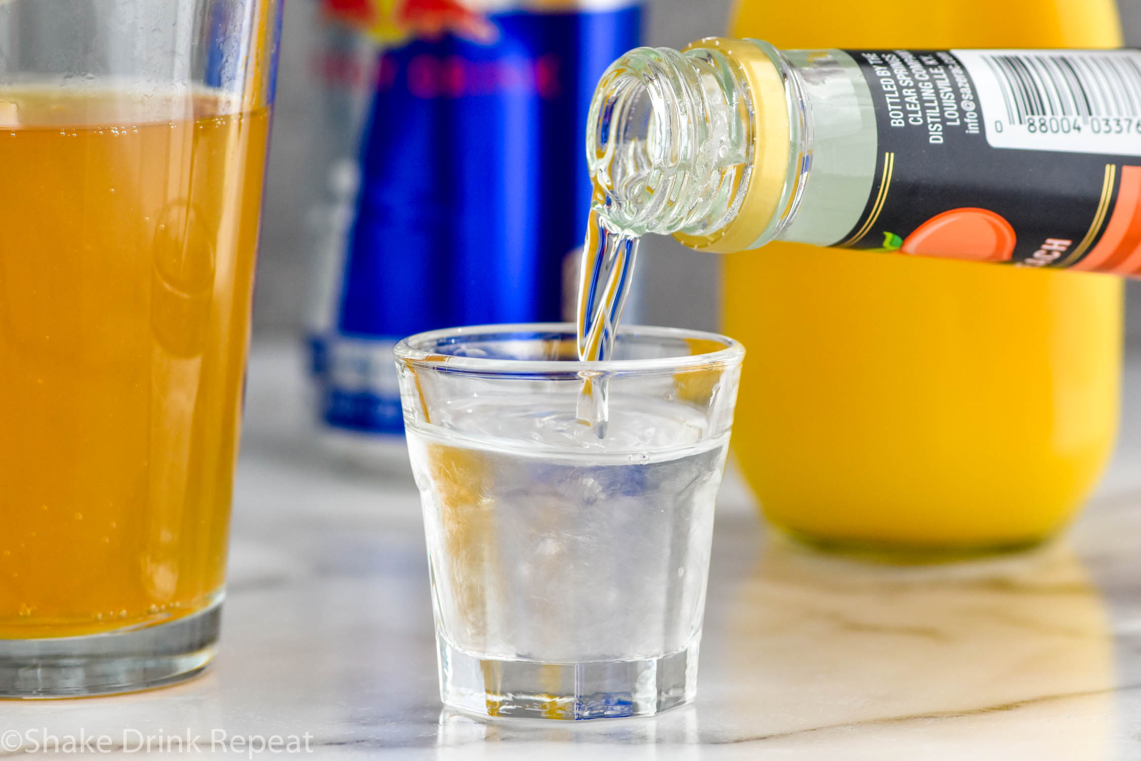 Side view of peach schnapps being poured into a shot glass for Cactus Cooler Shot recipe. Pint glass of Red Bull, can of Red Bull, and bottle of orange juice in the background.