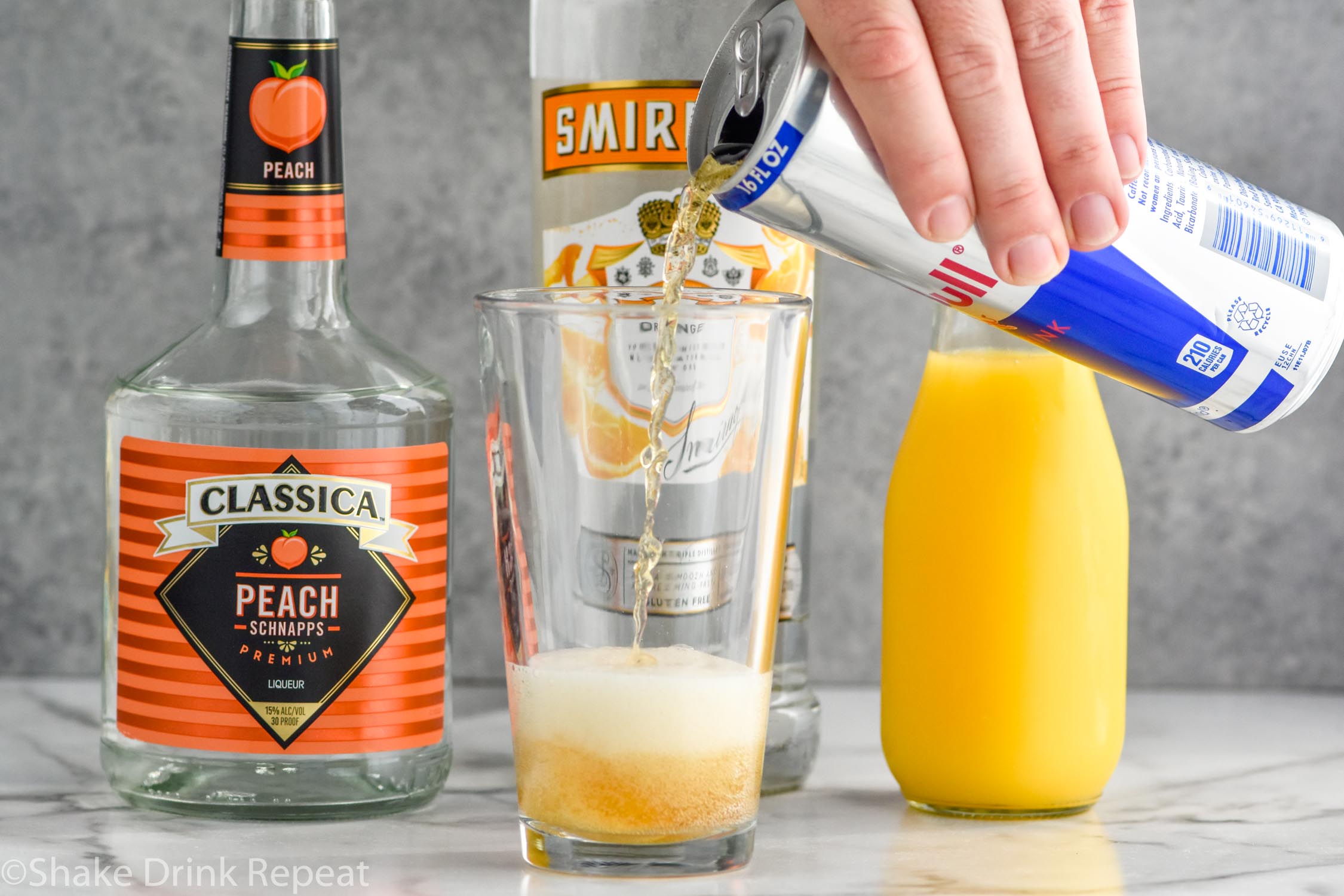 Side view of person's hand pouring can of Red Bull into a pint glass for Cactus Cooler Shot recipe. Bottles of peach schnapps, mandarin vodka, and orange juice behind glass.