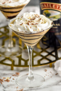 Baileys Martini topped with whipped cream, martini and bottle of baileys irish cream sitting in background.