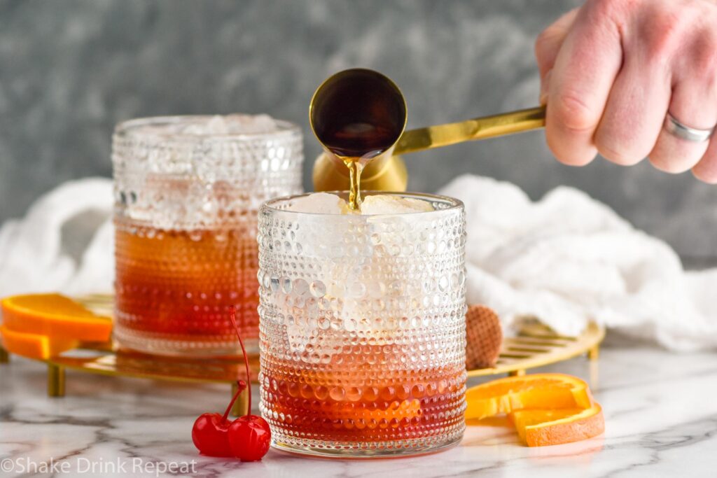 Side view of person's hand pouring cocktail jigger of ingredients into tumblers for Cherry Old Fashioned recipe. Orange slices and cherries beside.