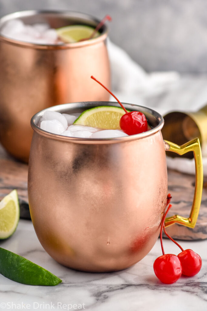 Cherry Moscow Mule served in copper mugs with ice, lime wedges, and cherries as garnish. More lime wedges and cherries on counter beside.