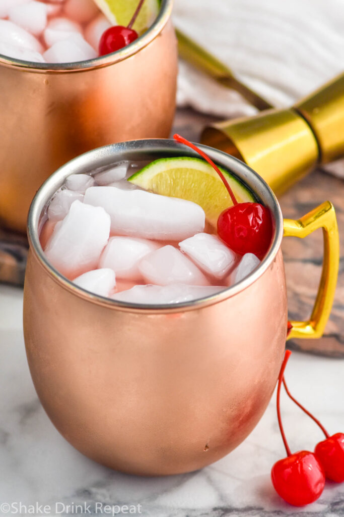 Cherry Moscow Mule served in copper mugs with ice, lime wedge, and cherry as garnish. Cocktail jigger and more cherries beside.