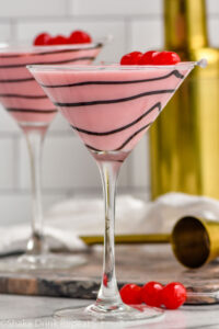 Side view of Chocolate Covered Cherry Martini with chocolate swirled on the glass and cherries as garnish. Cocktail jigger and cherries for garnish beside.