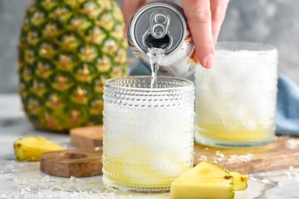 Man's hand pouring can of coconut soda into a glass of Pina Colada Vodka Soda ingredients and ice. Fresh pineapple and glass of pina colada vodka soda sitting in background