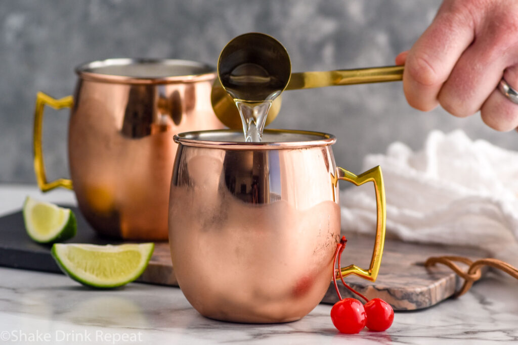 Side view of person's hand pouring cocktail jigger of ingredient into copper mugs for Cherry Moscow Mule recipe. Cherries and lime wedges beside for garnish.