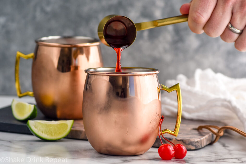 Side view of person's hand pouring cocktail jigger of grenadine into copper mugs for Cherry Moscow Mule recipe. Lime wedges and cherries beside for garnish.