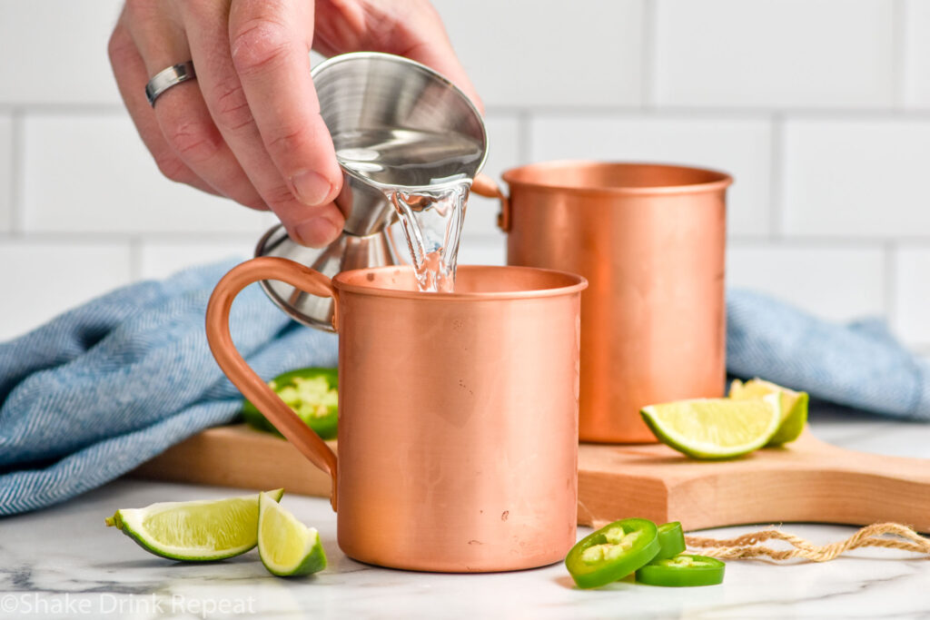 Side view of person's hand pouring vodka into copper mugs for Jalapeno Moscow Mule recipe. Lime wedges and sliced jalapenos beside as garnish.