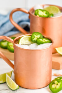Jalapeno Moscow Mules served in copper mugs with ice, sliced jalapenos, and lime wedge as garnish. More sliced jalapenos and lime wedges beside copper mugs.