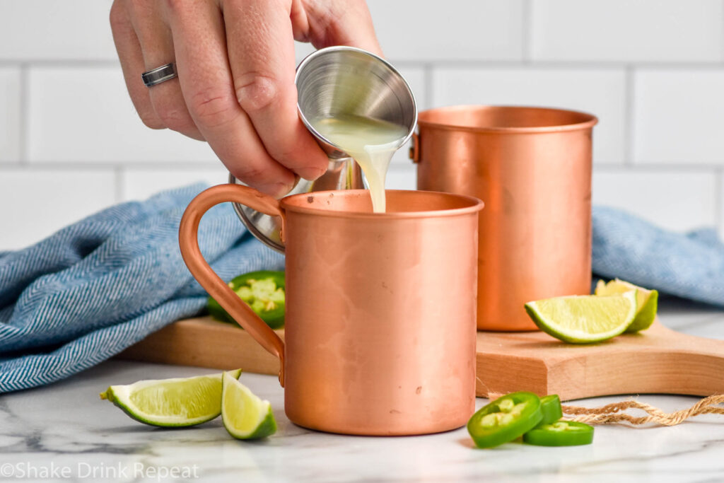 Side view of person's hand pouring ingredient into copper mugs for Jalapeno Moscow Mule recipe. Lime wedges and sliced jalapenos beside mugs for garnish.