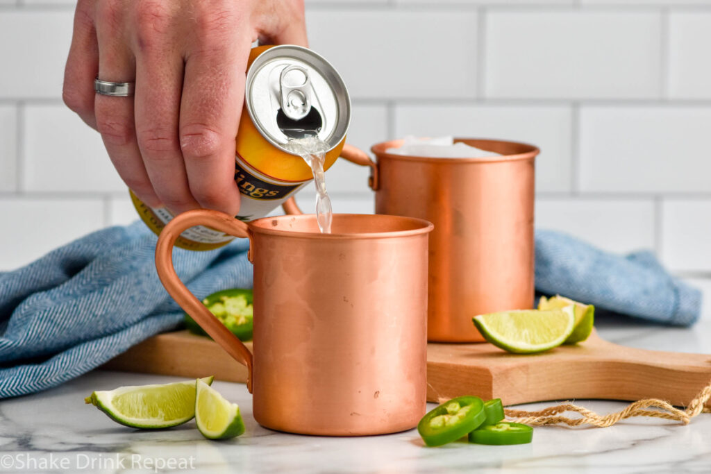 Side view of person's hand pouring can of ginger beer into copper mug for Jalapeno Moscow Mule recipe. Lime wedges and sliced jalapeno beside copper mugs as garnish.