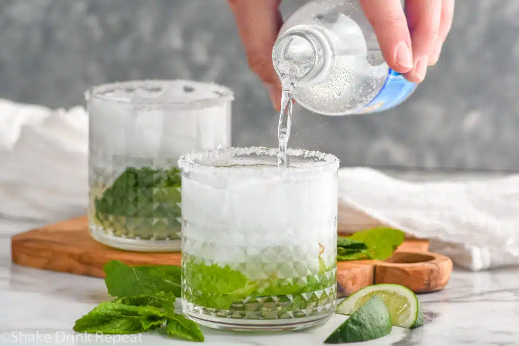 man's hand pouring bottle of club soda into a glass of mojito margarita ingredients and ice. Glass of mojito margarita, fresh mint leaves, and lime wedges sitting in background.