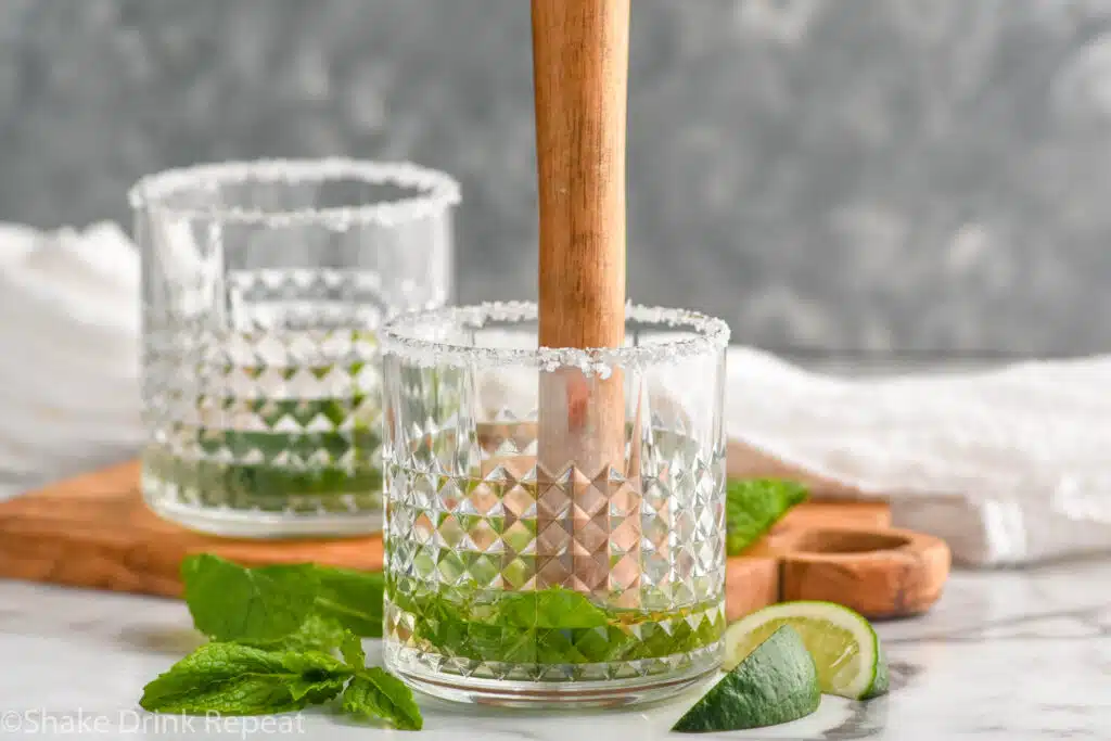 muddler muddling Mojito Margarita ingredients in a glass. Glass, fresh mint leaves, and lime wedges surrounding.