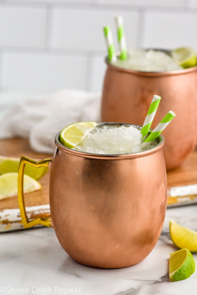Moscow Mule Slush served in copper mugs with straws and lime wedges. Lime wedges beside for garnish.