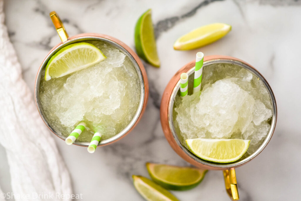 Overhead view of Moscow Mule Slush served in copper mugs with straws and lime wedges. Lime wedges beside copper mugs for more garnish.