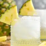 two glasses of Pina Colada Vodka Soda with ice and garnished with pineapple wedge. Fresh pineapple sitting in background.