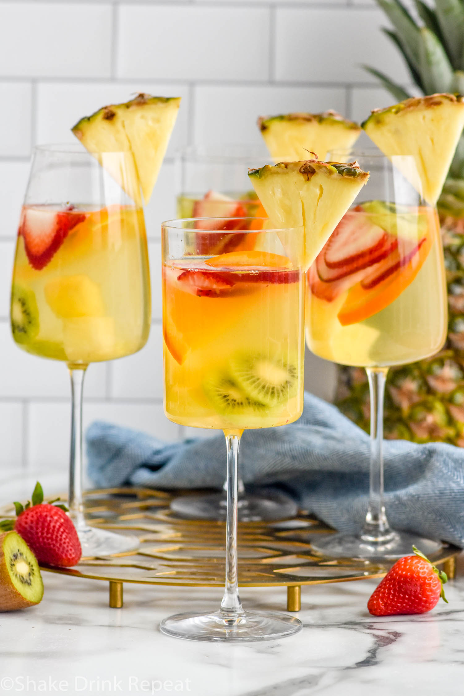 Four wine glasses of Tropical Margarita Sangria garnished with a slice of pineapple. Fresh strawberries and kiwi sitting beside.