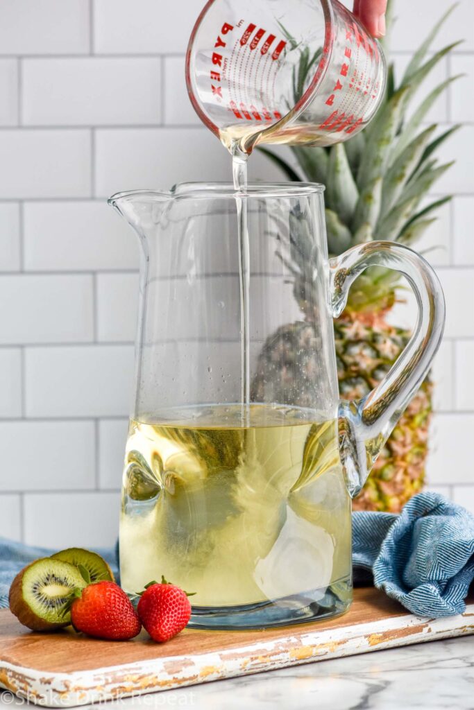 Glass measuring cup of liquid being poured into pitcher of ingredients for Tropical Margarita Sangria recipe. Kiwi, strawberries, and whole pineapple beside.